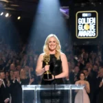 Amy Poehler clutching her Golden Globe at podium with spotlight and Good Hang logo behind her