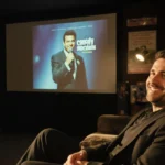 André Ricciardi smiling warmly on worn couch with comedy memorabilia and colonoscopy machine in background