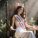 Andrea Andrade sits on worn wooden throne with crown and sash beside her surrounded by lush greenery