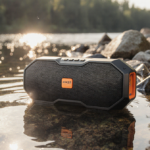 Anker Bluetooth speaker rests partially submerged in a lake shoreline with warm sunlight reflecting and lush rocks nearby