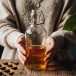 Person holding apple cider vinegar bottle with candle light and green leaves around a rustic wooden table.