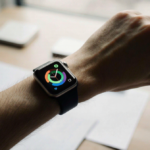Wrist wearing an Apple Watch illuminated by soft natural light with a blurred neutral desk and scattered documents nearby.