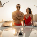 Ashton Kutcher posing in red swimsuit with Mila Kunis beside him and golden sunset over luxury speedboat