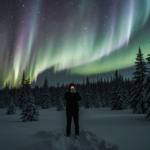 Figure standing with hands cupped around mouth with aurora lighting snow-covered evergreen forest behind dark sky