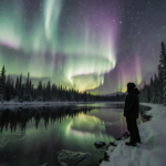 Silhouetted figure gazing up at shimmering aurora with snow‑covered trees and frozen lake reflecting the lights.