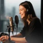 A young woman is speaking into a microphone with headphones on near a wooden desk.