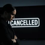 Meghan Markle stands alone in front of a dimly lit black screen displaying