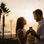 Groom proposing with diamond ring box as bride-to-be cries tears of joy near rebuilt palm trees with sunset beach and wildfir