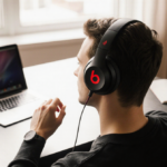 Person examining Beats Studio Pro headphones with laptop and coffee on a minimalist desk under warm window light