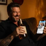 Blake Shelton sits on worn couch with whiskey bottle and phone showing tabloid article about Gwen Stefani divorce