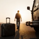 Young family member carrying backpack walks toward Mexico border with old suitcase and van at sunrise
