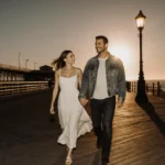 Bradley Cooper and Gigi Hadid walking hand in hand on Santa Monica pier at sunset with golden light and vintage streetlamp