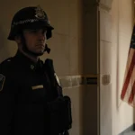 Capitol Police officer stands alone in dim hallway with faded flag and warm light and askew helmet and tired eyes.