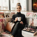 Catherine Santino smiles while holding designer handbag and coffee with fashion magazines and makeup station in bright coffee