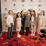 Celebrity kids laughing and posing on red carpet with paparazzi cameras and designer bags scattered in front