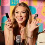 Céline Dion playfully holds lip gloss and microphone with colorful eclectic background behind her