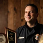 Chief Sam DiGiovanna smiles softly in fire uniform with awards and warm wooden background behind him