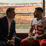 Clark Hunt leaning forward to talk with Travis Kelce in golden light with Arrowhead Stadium windows reflecting autumn leaves
