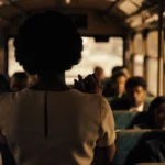 Claudette Colvin stands defiantly on a 1950s bus with shadowy passengers seated behind her and city lights glowing through th