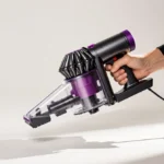 Hands holding cordless vacuum with cord wrapped around handle and foot shadow behind on white background
