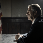 Somber middle-aged man sitting at metal table with a faded flag and faint spotlight in a courtroom.