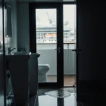 Cruise ship bathroom reflects sink and toilet in puddle with eerie glow seeping from door.