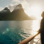 A lone passenger stands at the cruise ship railing with golden sunset light and Marquesas Islands mountains rising through mi