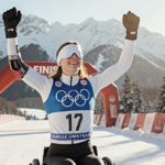 Danelle Umstead raising arms in triumph at Paralympic ski finish line with blindfold and prosthetic leg against snowy backdro