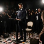 David Beckham stands at podium with empty chair and paparazzi flash in background