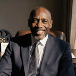 Delroy Lindo sits at a desk wearing a navy suit with a tie smiles at the camera with a blurred film reel in the background.