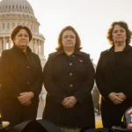 Three grieving widows clasp hands with firefighter helmets and black ribbons at their feet in front of Iowa State Capitol