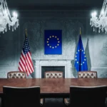 Empty chairs face each other across a wooden table with American and EU flags showing frosty patterns