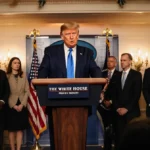 Donald Trump speaking at the podium during a White House press briefing with journalists lined up and flag in background