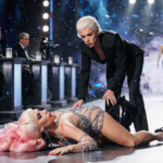 RuPaul contestant lying on runway with Law Roach standing over them looking critical amid blurred judges tables and cameras