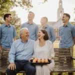 Mark and Aimee Maye laugh on a wooden bench with their four sons standing behind them near UNC campus