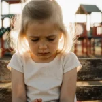Young Drew Barrymore sits alone on wooden bench with head down and playground blurred behind her