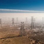Power lines stretch across cracked desert land with heat waves distorting towers under blue sky