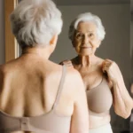 Elderly woman adjusting wireless bra straps while checking her reflection in mirror with natural light illuminating her hair