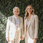 Ellen DeGeneres and Portia de Rossi holding hands with smiles in front of lush greenery and birthday balloons.