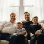 Elton John and David Furnish sitting with their children on a sunlit couch with colorful artwork and family photos visible be