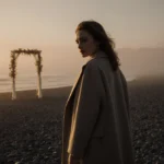 Emeli Sandé stands alone on a beach with her back to the viewer and a distant wedding arch visible through the mist