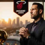 Erik Spoelstra stands confidently with hands clasped in front of Miami Heat championship banner and sunset skyline