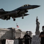 F-16 hovering above Venezuelan presidential palace with US flag on landing gear and protestors gathering at front.