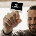 Father holding newborn baby hand with black-and-white polaroid caption Tag You