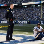 FBI agent standing beside yellow crime scene tape with somber T.Y. Hilton seated in shade on Indianapolis Speedway infield.