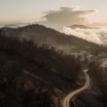 Winding dirt road disappears into haze with fog swirling at mountain base and golden glow over horizon