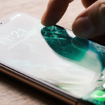 Person’s hand reaching to adjust settings with iPhone screen showing locked Liquid Glass home under warm lighting.