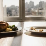 Split table shows full plate of roast chicken and vegetables facing empty plate with faded food remains while sunlight stream