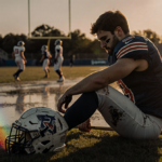 Jake Eldridge sits alone on a wet football field with a baseball bat and reflective stadium lights