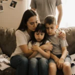 Foster mom embraces two children on worn couch with scattered papers and photos showing hope